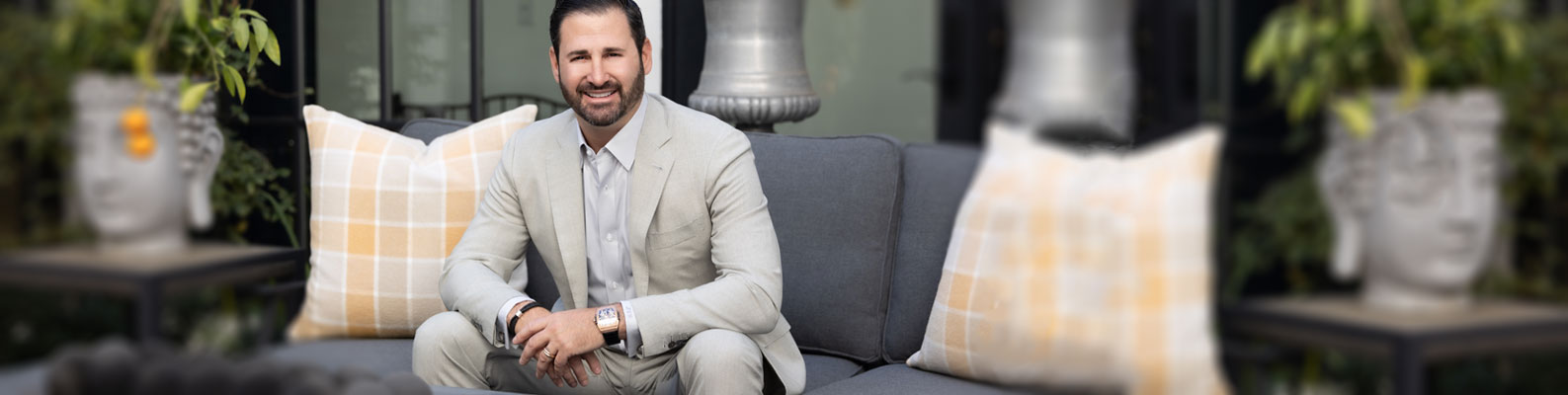 Dave Cantin, business finance speaking professional, wearing a tan suit posing for a photo on a gray couch with yellow plaid pillows and two planters on the side.