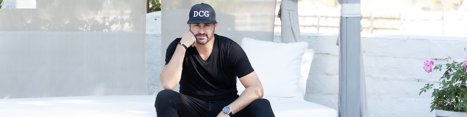 Dave Cantin, a business speaker, wearing an all-black outfit and a black DCG hat, posing on a white sofa.